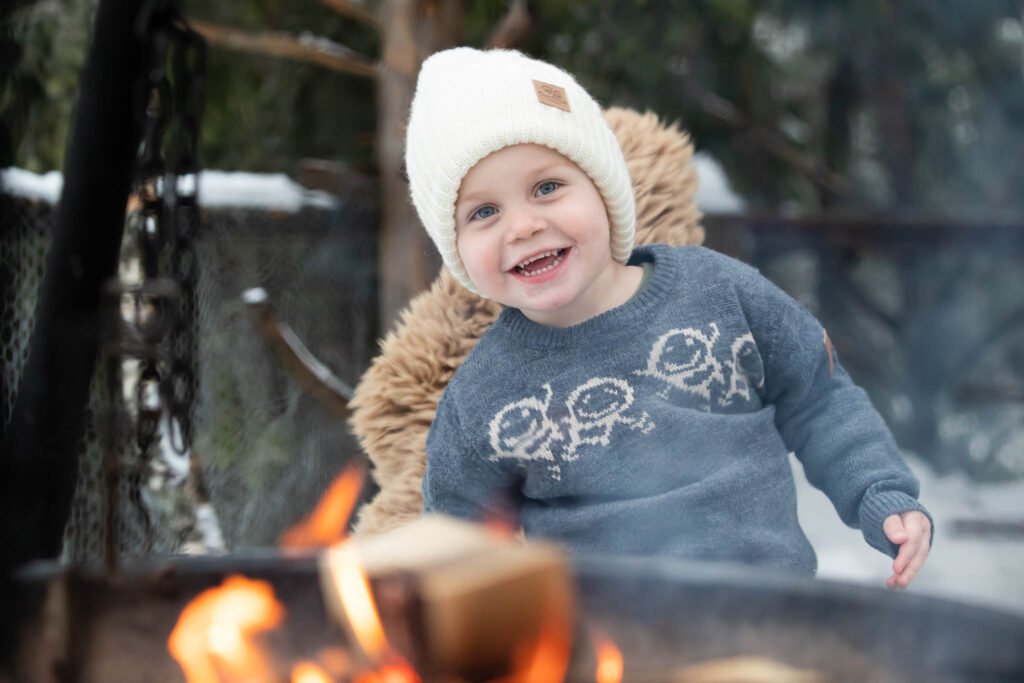 Liten gutt som ler og koser seg ved en bålpanne ute om vinteren. Husk å gjør vinterturen til gode minner.